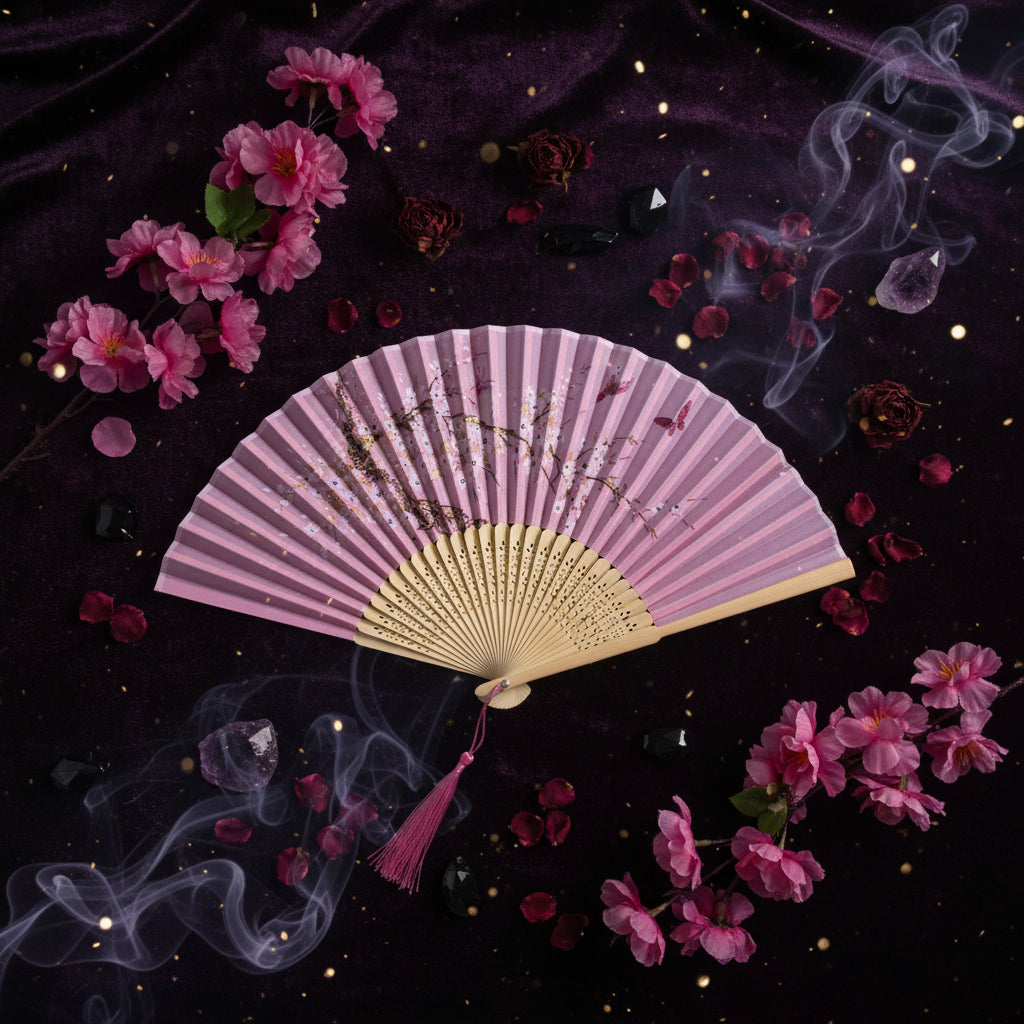 A pink folding hand fan with delicate floral and branch illustrations, displayed on a white fabric background with pink cherry blossoms and a pink tassel.