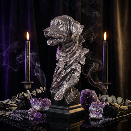 A metallic bronze dog bust sculpture on a black pedestal base, displayed on a wooden surface with a blurred sofa in the background.