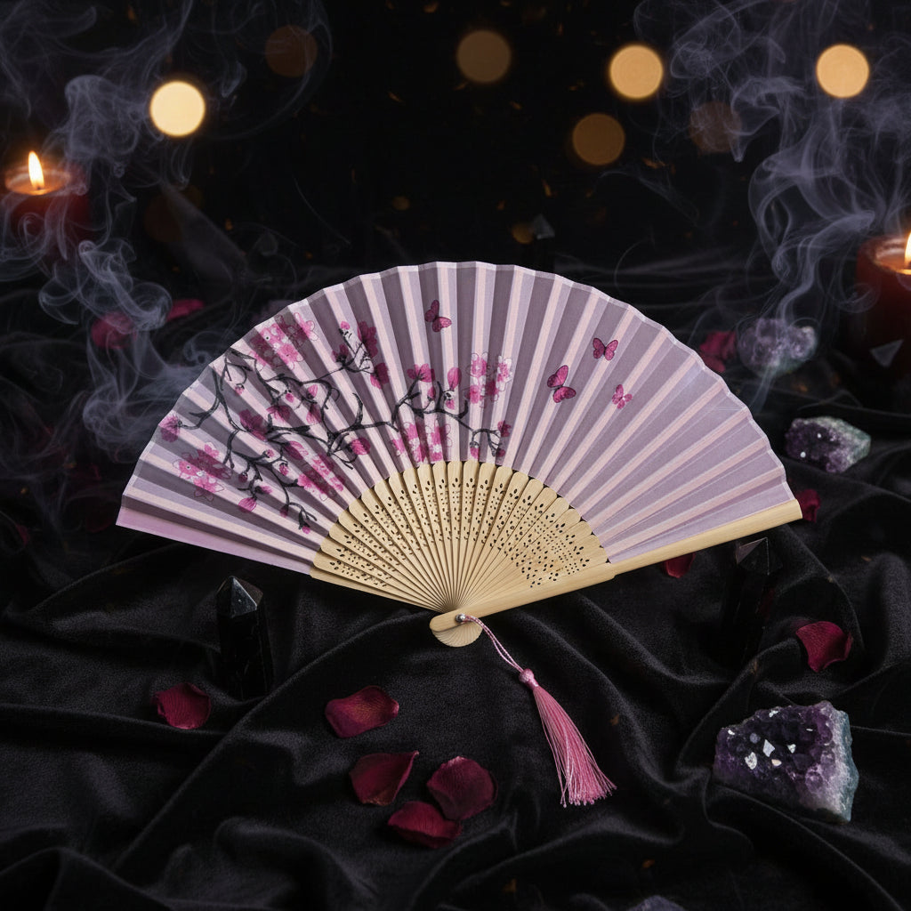 A traditional folding hand fan with a light pink and white patterned paper canopy, printed with black branches, pink blossoms, and butterflies. The fan has a beige wooden frame and a pink tassel, styled with pink cherry blossoms on a white background.