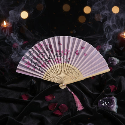 A traditional folding hand fan with a light pink and white patterned paper canopy, printed with black branches, pink blossoms, and butterflies. The fan has a beige wooden frame and a pink tassel, styled with pink cherry blossoms on a white background.