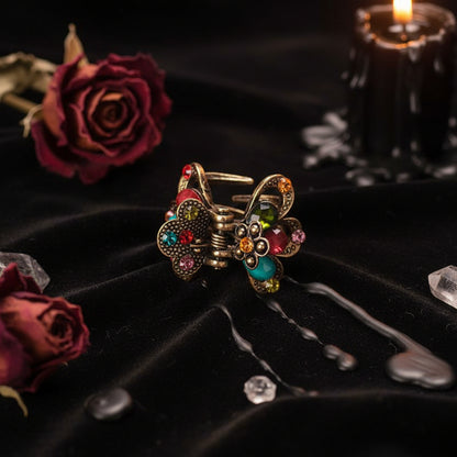 A vintage-style butterfly hair claw clip with an antique bronze metal body and colorful rhinestone and bead accents in red, turquoise, green, orange, and pink, resting on a textured white fabric background.