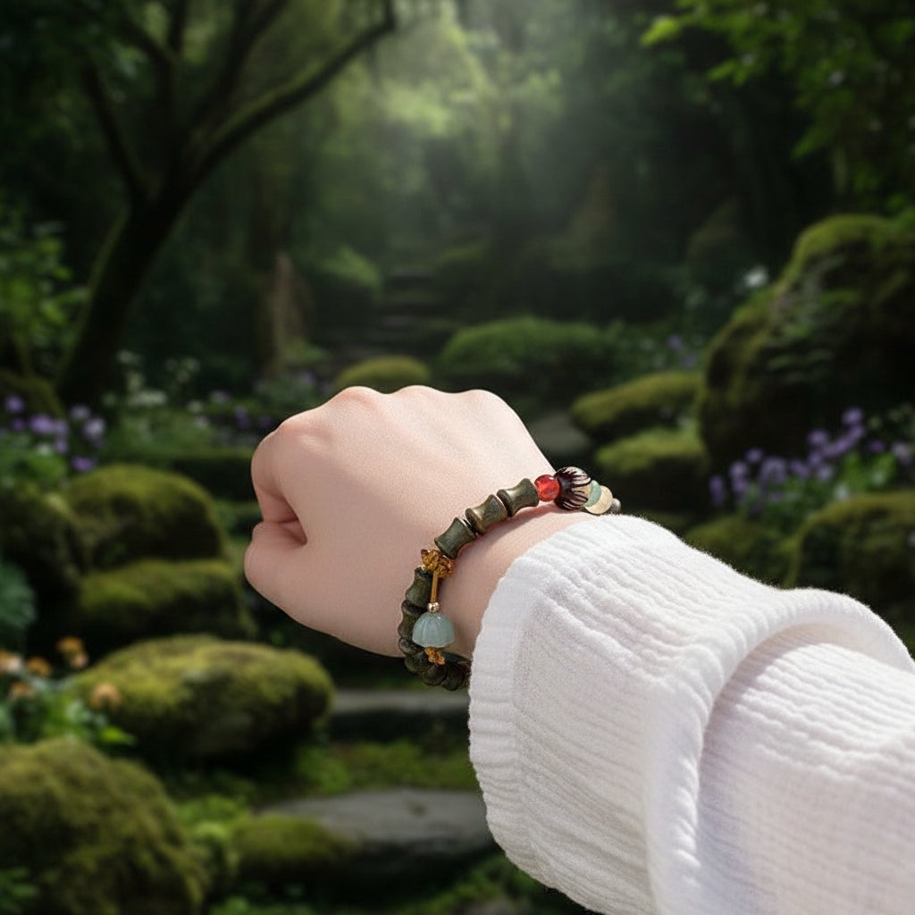 A close-up of a person's wrist wearing a beaded bracelet with bamboo-style green beads, a small red bead, a black-and-white patterned accent, and a dangling pale green bell charm, set against a soft white textured garment.