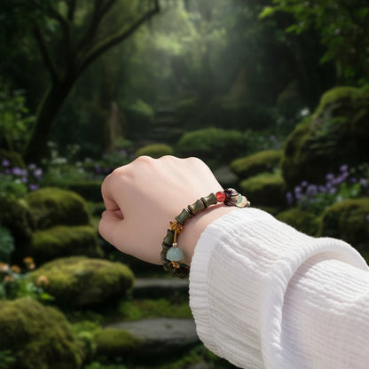 A close-up of a person's wrist wearing a beaded bracelet with bamboo-style green beads, a small red bead, a black-and-white patterned accent, and a dangling pale green bell charm, set against a soft white textured garment.