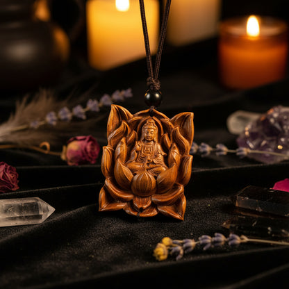 A close-up of a carved wooden pendant depicting a seated Buddha figure surrounded by lotus petals, hanging from black beads on a string. The pendant is shown resting on a wooden surface with a dark bowl and dried leaves in the background.